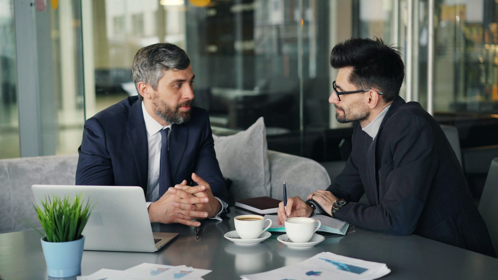 Two business leaders in suits in a one-to-one conversation over coffee at an office table, one listening closely as the other speaks.