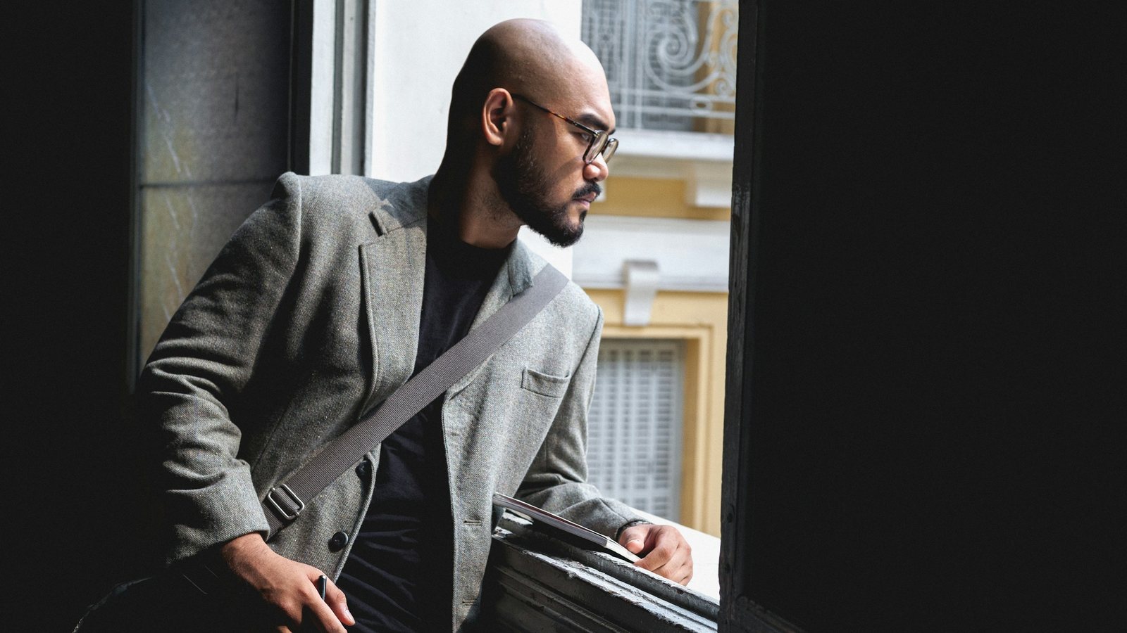 A leader in a grey jacket leaning by an open window, looking out and reflecting, a picture of the honest thinking good coaching makes room for.